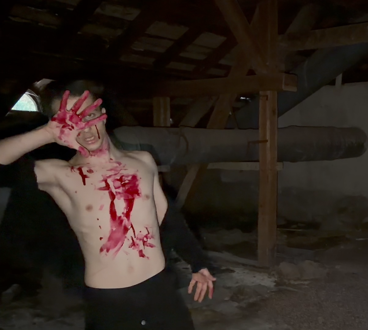 A blood-covered figure raising a red-stained palm toward the camera in a dark attic